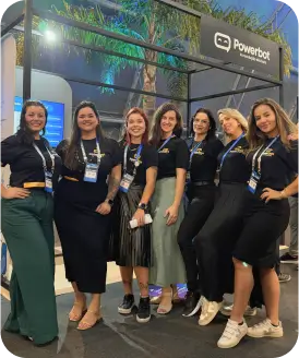 Grupo de mulheres em evento da Powerbot, todas com camisetas escuras e poses descontraídas, representando diversidade e empoderamento feminino.